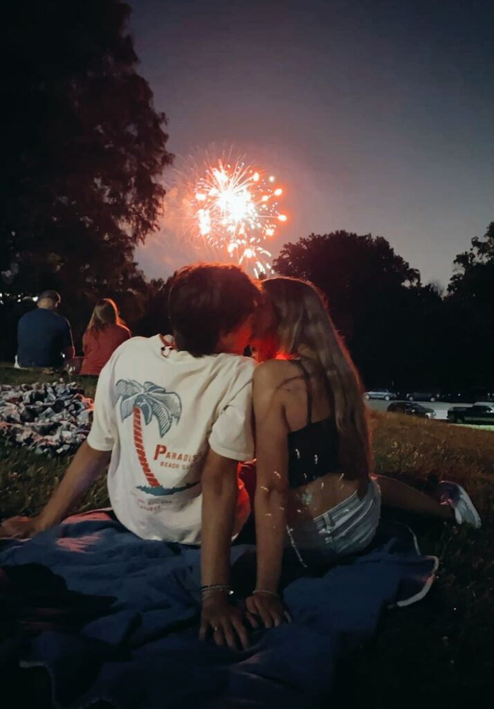 Couple watching fireworks on a blanket in a park at night, sky illuminated by colorful bursts.