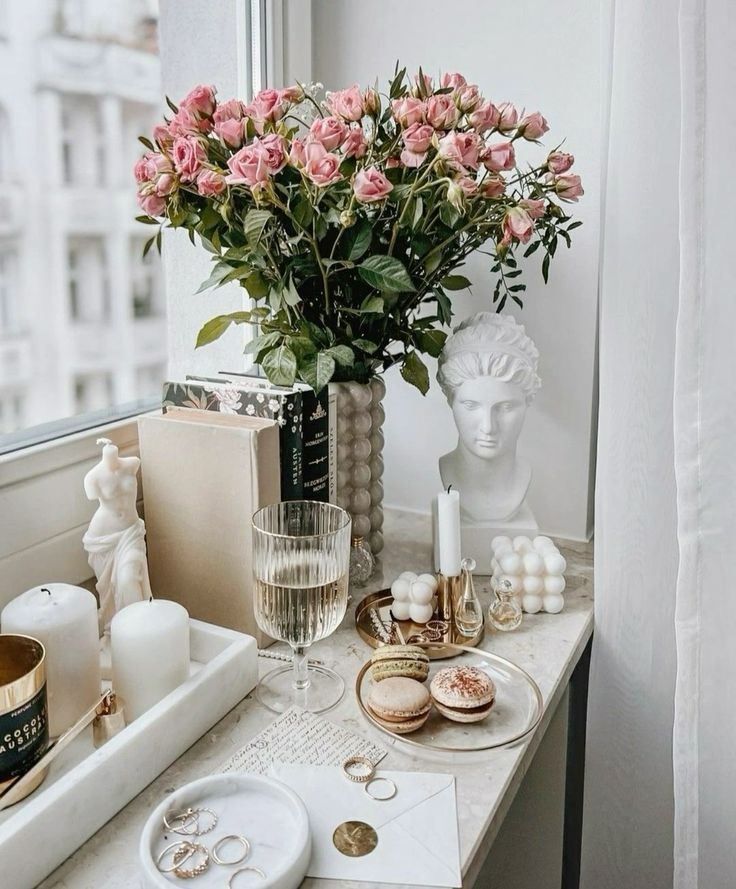 Elegant room decor with pink roses, candles, gold accents, a wine glass, and macarons on a marble table.