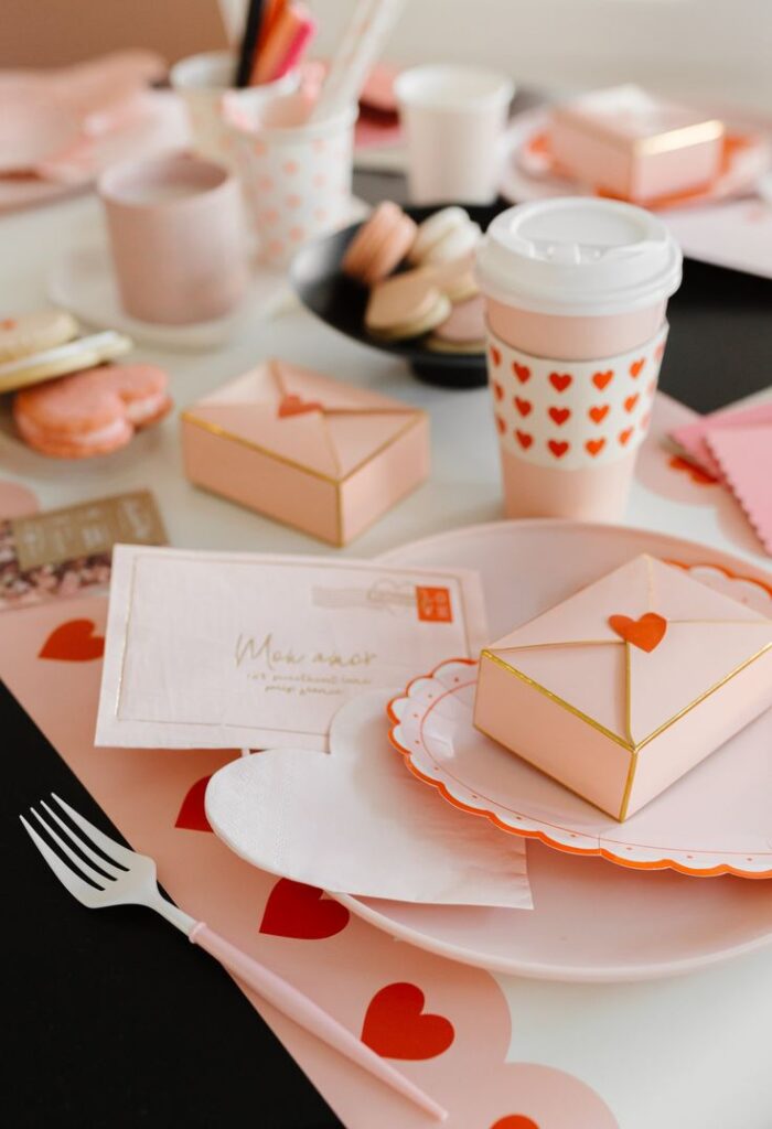 Romantic themed setup with heart-patterned coffee cup, gift boxes, macarons, and pink envelopes on a table.