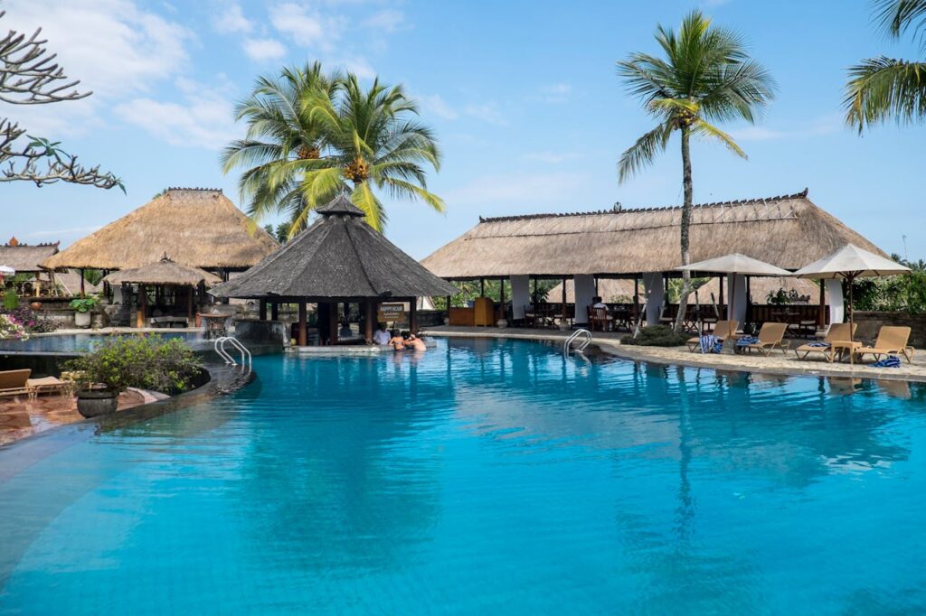 Tropical resort pool with thatched-roof bar and palm trees under clear blue sky, perfect for relaxing vacation.