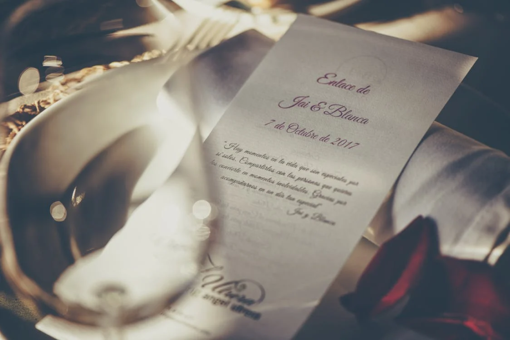 Elegant wedding invitation with script text on a dining table, surrounded by glassware and a rose petal.