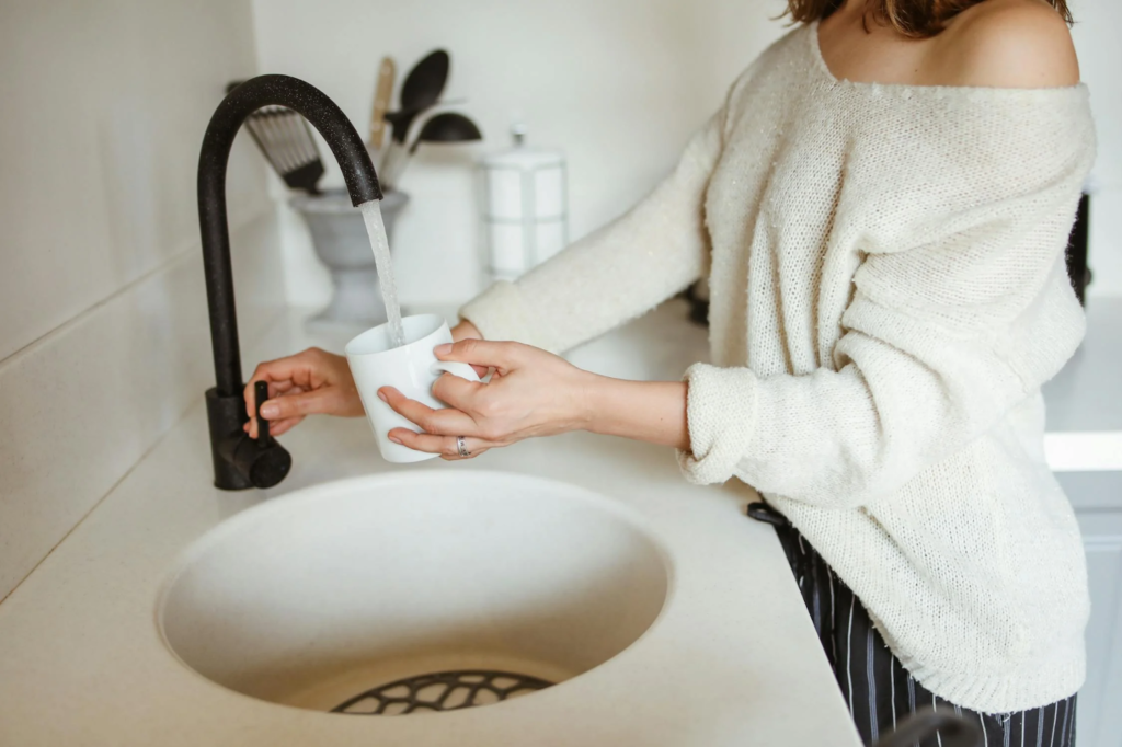 Woman filling white mug with water from modern kitchen faucet.