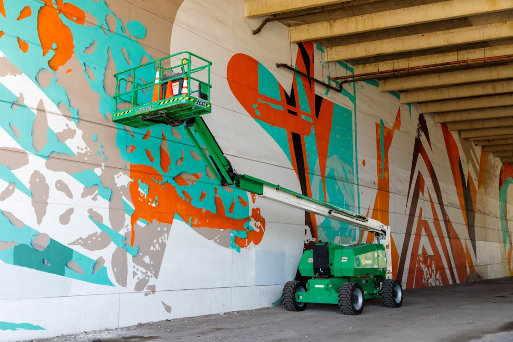 Boom lift painting vibrant mural on concrete wall under bridge.