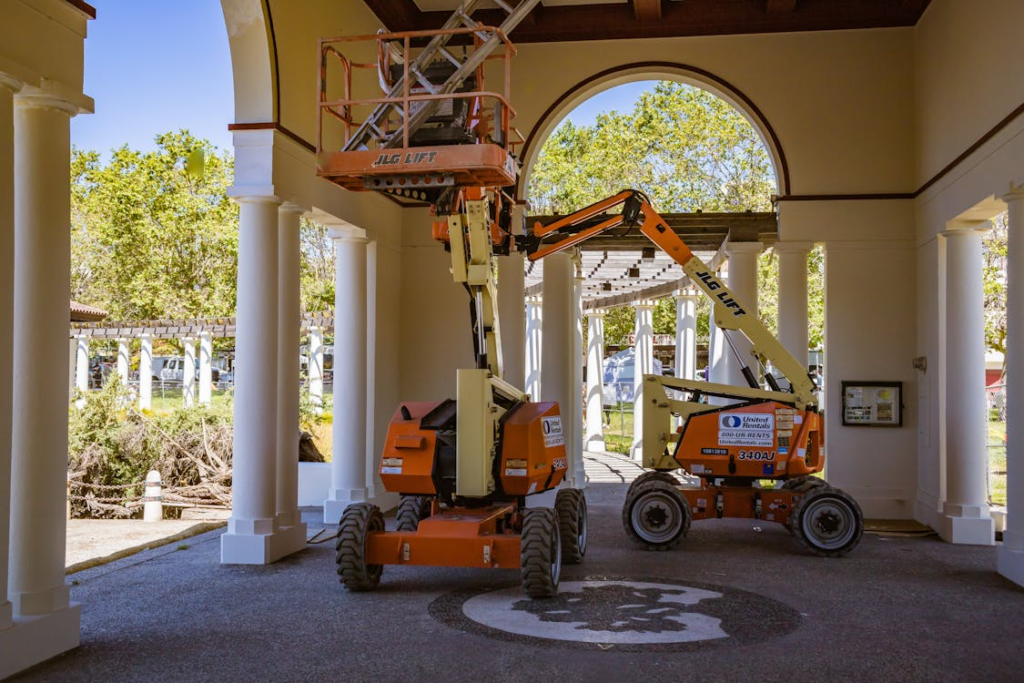 Orange lift equipment under an arch in a sunny outdoor setting with white columns and trees.