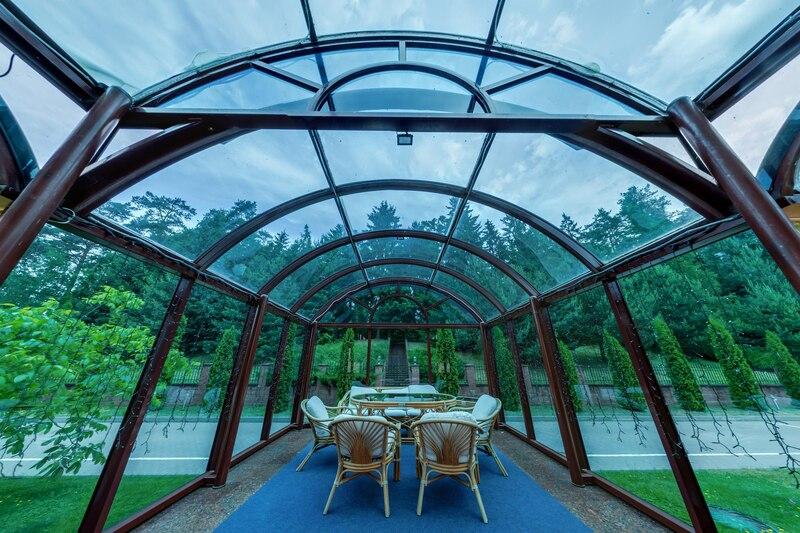 Glass-enclosed outdoor seating area with lush greenery background and stylish rattan chairs under a clear blue sky.