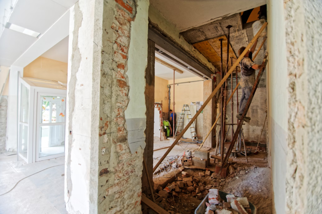 Construction worker renovating building interior, using scaffolding and tools amidst exposed brick and debris.