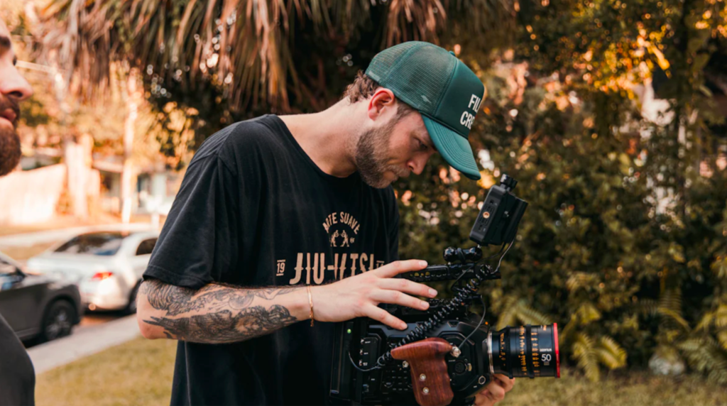 Man with tattoos and green cap operating professional camera outdoors.