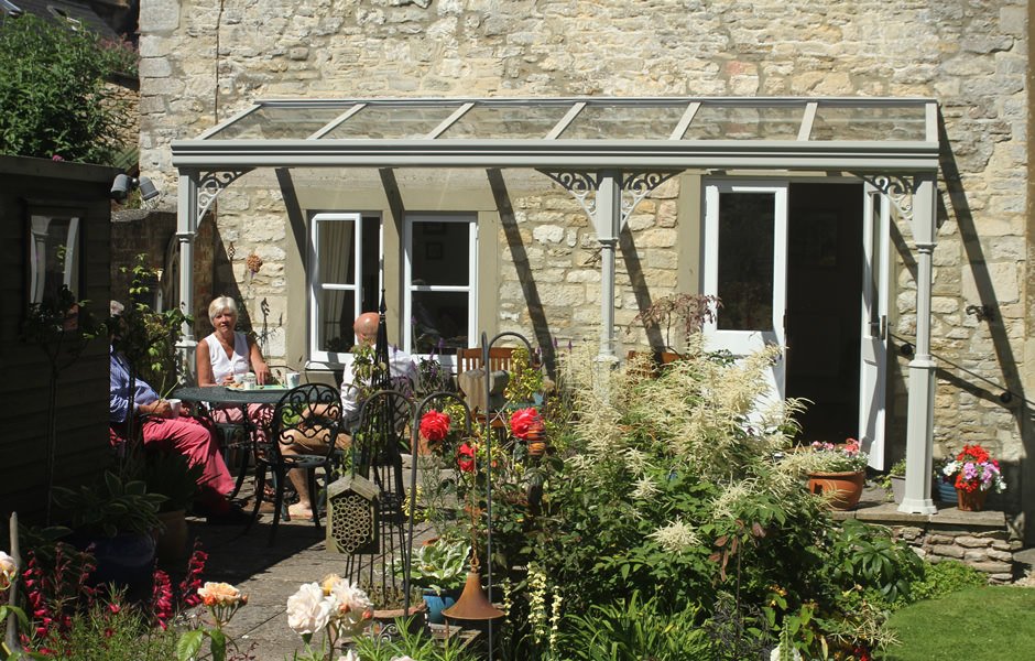 Garden patio with people sitting under a glass canopy, surrounded by colorful flowers and rustic stone wall.