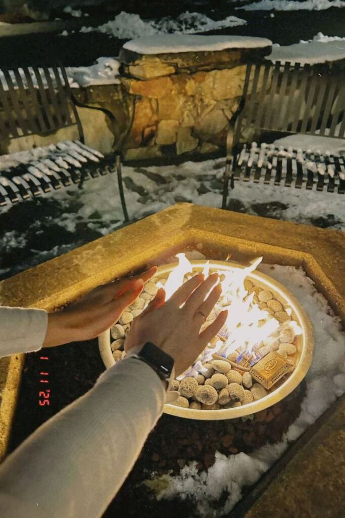 Hands warming by fire pit in snowy outdoor setting, night warmth and cozy winter atmosphere.