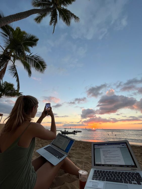 Woman working on laptops at a tropical beach during sunset, capturing the view with her phone.