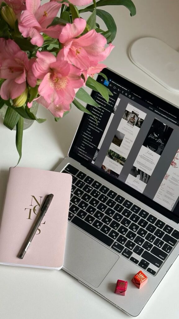 Laptop with graphic design software, pink flowers, a notebook, pen, and chocolates on a white desk.