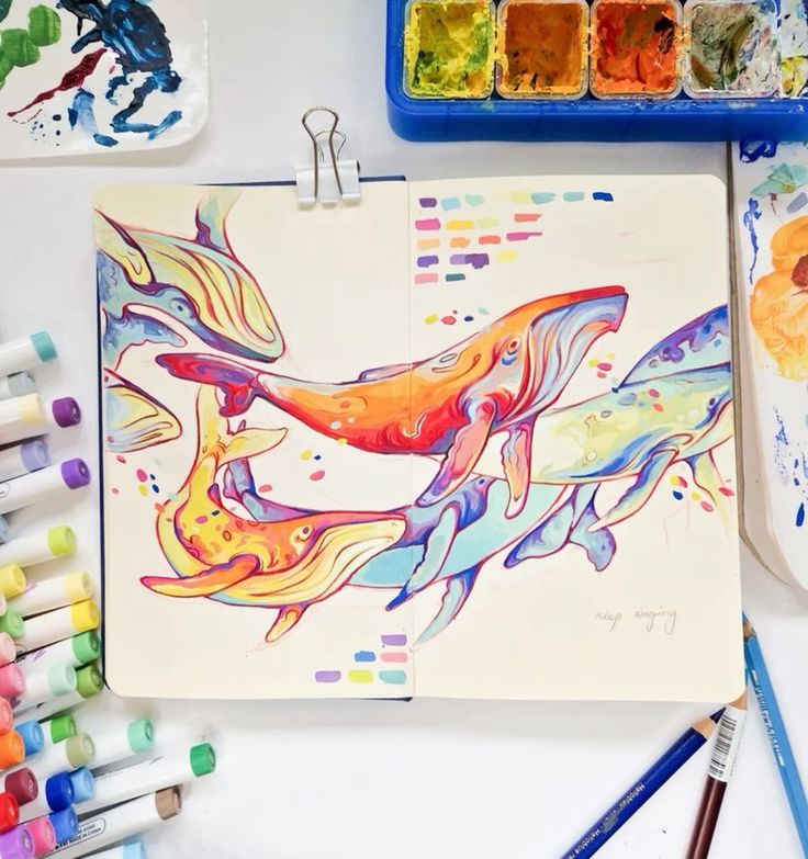 Colorful whale illustration in sketchbook with art supplies around it.