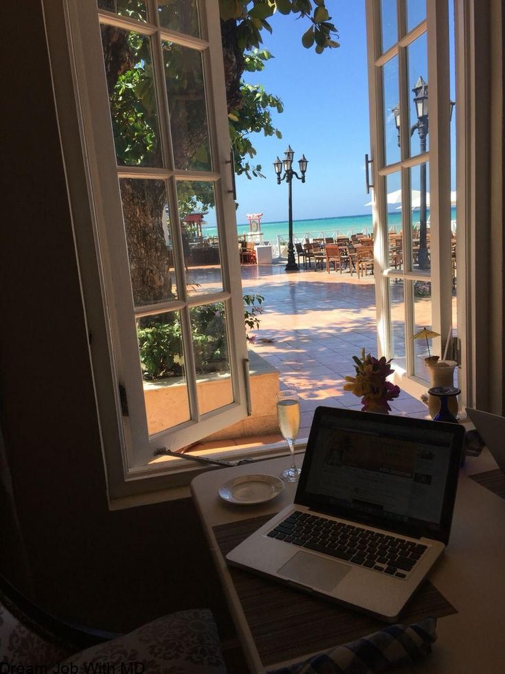 Open window view of oceanfront café table with laptop, champagne, and sea breeze under sunny skies.