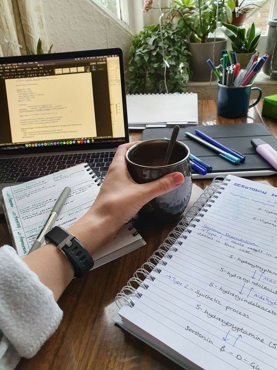 Person holding a coffee mug at a study desk with a laptop and open notebooks, surrounded by pens and notes.