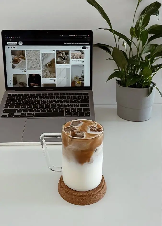 Iced coffee on coaster, laptop with Pinterest, and houseplant on a white desk for a cozy workspace vibe.