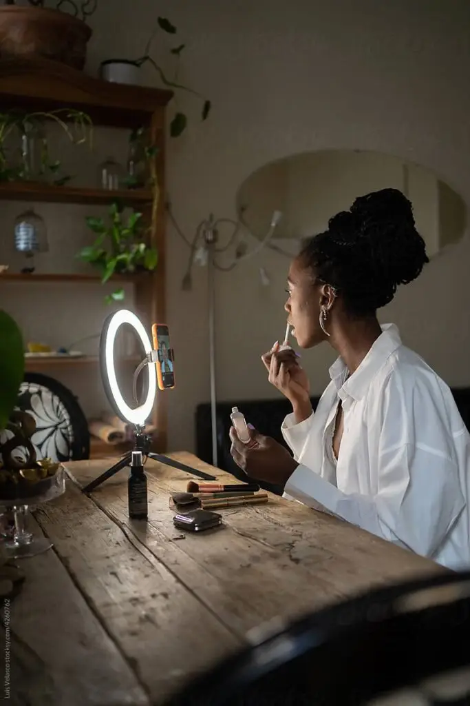 Woman applying makeup with ring light and smartphone on a wooden table.