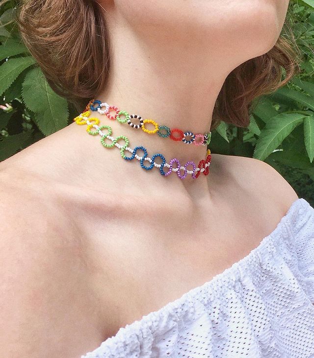 Woman wearing colorful beaded choker necklaces, with a white off-shoulder top, in a leafy outdoor setting.