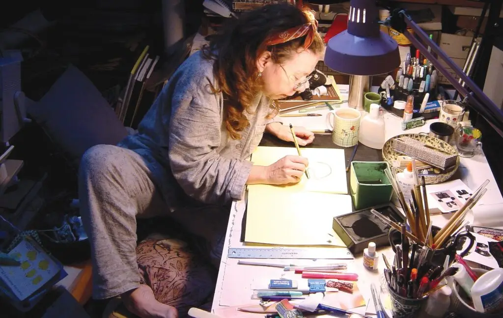 Artist drawing at a cluttered desk with various art supplies, cups, and a lamp illuminating the workspace.
