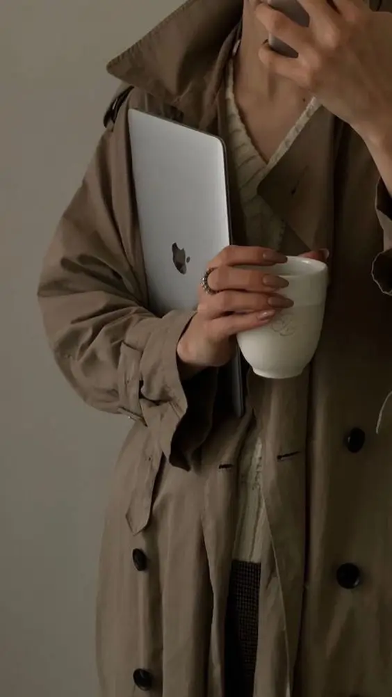 A person in a trench coat holding a laptop and a coffee cup, capturing a cozy and productive vibe.