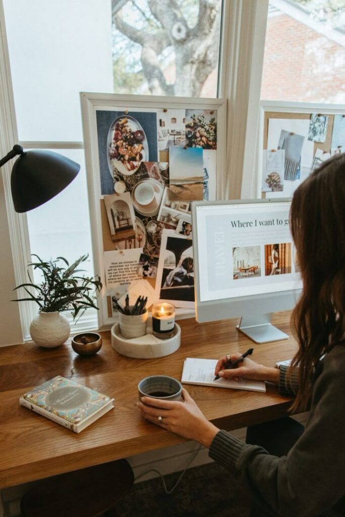 Woman writing at cozy desk with computer, mood board, coffee, and a candle.