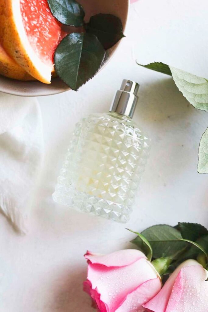 Perfume bottle with textured glass, surrounded by pink roses and fresh grapefruit on a white surface.