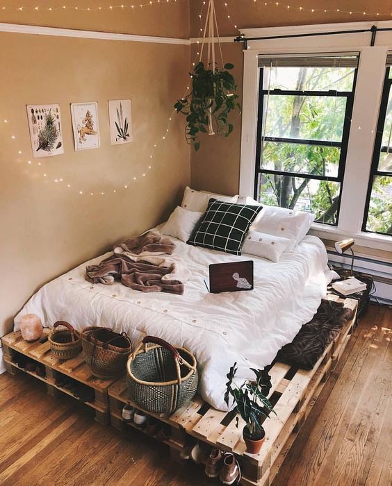 Cozy bedroom with pallet bed, string lights, plants, and natural decor by a window. Warm and inviting atmosphere.