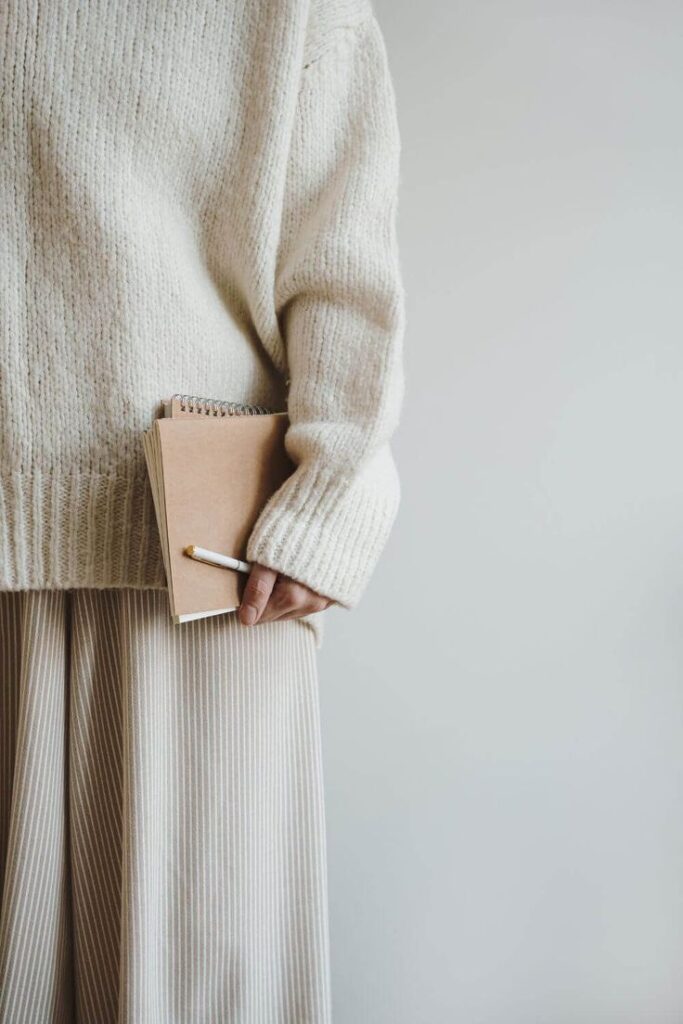 Person wearing a cozy sweater holds a notebook and pencil, showcasing minimalist fashion and creative inspiration.