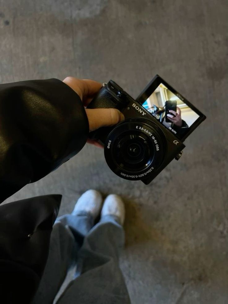 Person holding a Sony camera with a flip screen displaying their reflection, wearing casual shoes and jeans.