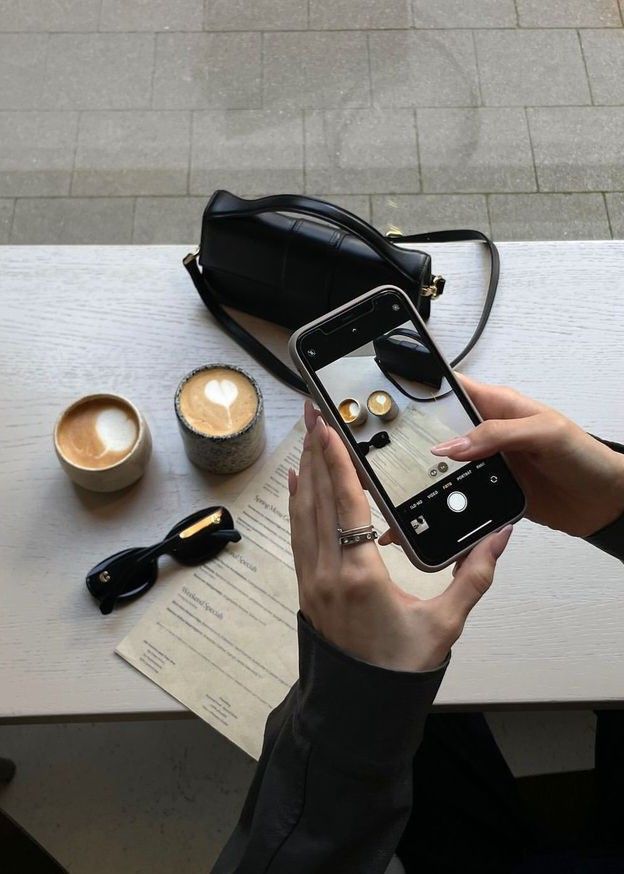 Person photographing coffee cups and sunglasses on a table with a smartphone, next to a menu and handbag.