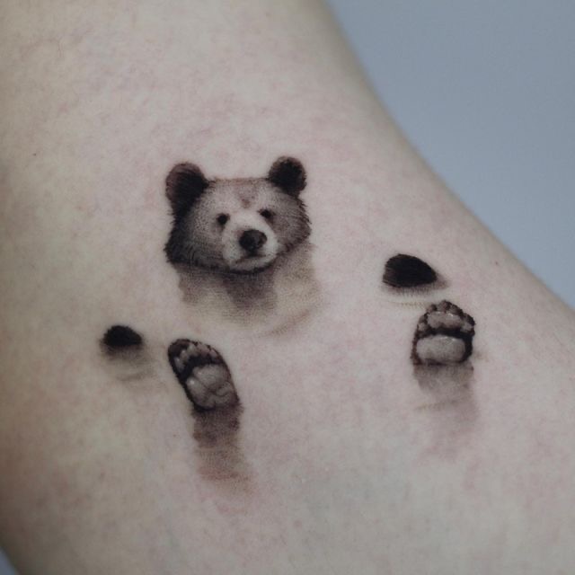 Tattoo of a cute bear peeking out, with its paws visible, on arm skin background.