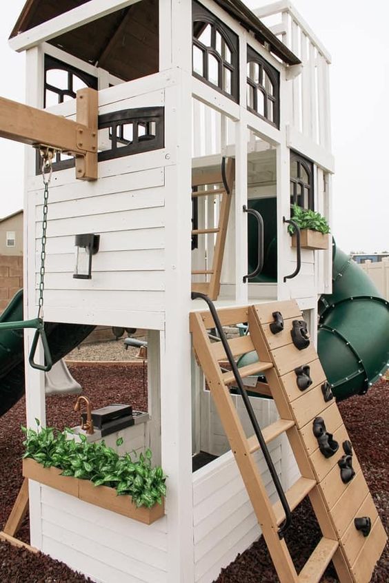 White wooden playhouse with a climbing wall, slide, and swing set in a backyard playground setting.