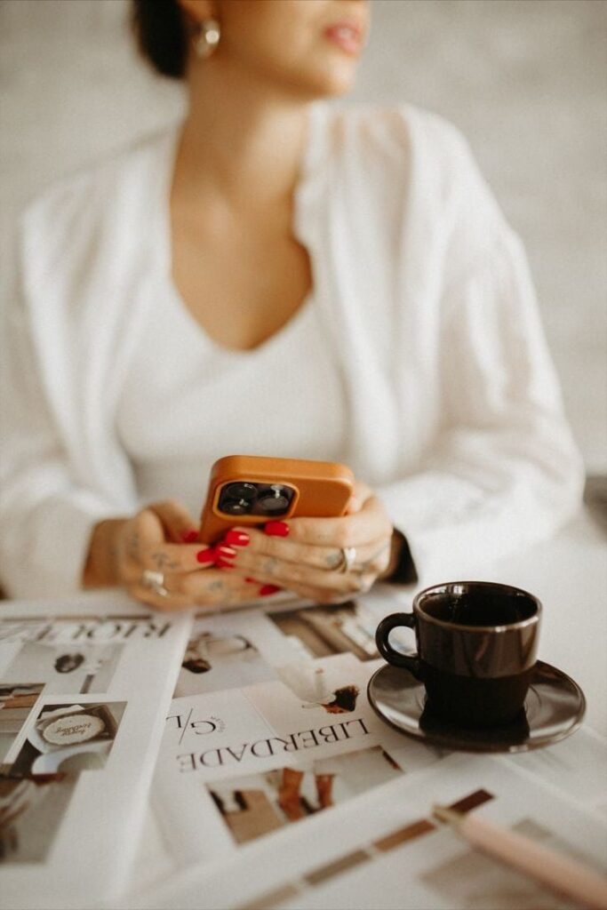 Woman with smartphone and coffee browsing fashion magazines at cozy table.