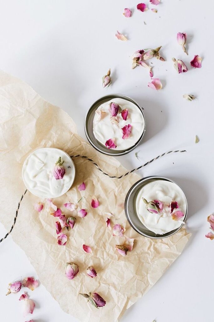 Three skincare cream jars with rose petals on parchment paper for a natural beauty treatment.