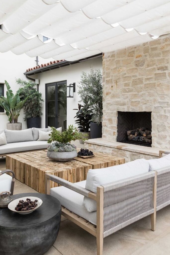 Modern outdoor patio with cozy seating, wooden table, stone fireplace, and plants under white canopy.