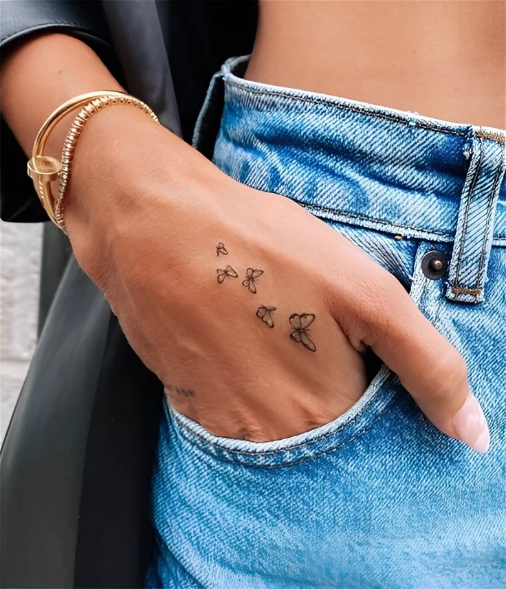 Hand with butterfly tattoos in pocket, wearing a gold bangle and blue jeans. Fashionable casual style.