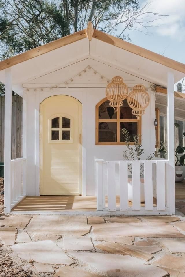 Charming wooden cottage with a yellow door and white porch, surrounded by trees and lit by woven lanterns.