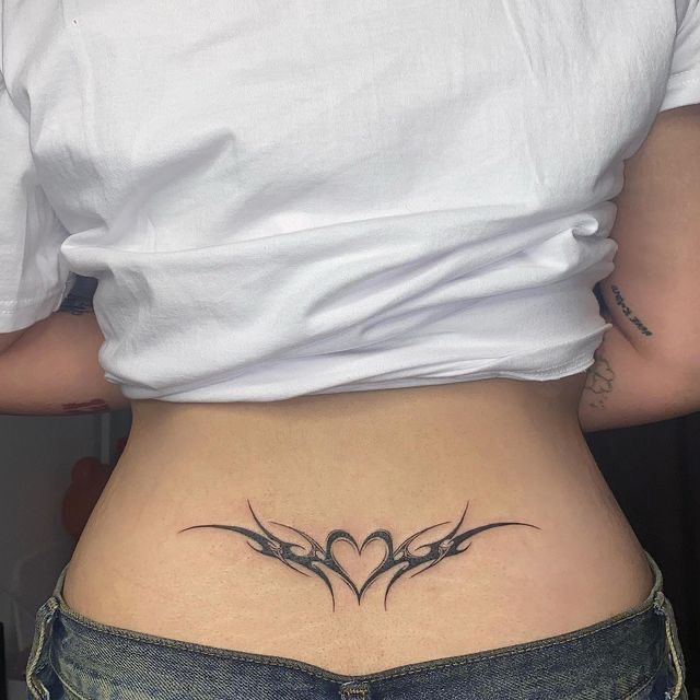 Lower back tattoo with tribal heart design, worn with a white top and jeans for a stylish look.