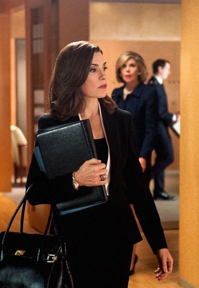 A professional woman in an office hallway carrying a leather folder and handbag, with colleagues in the background.