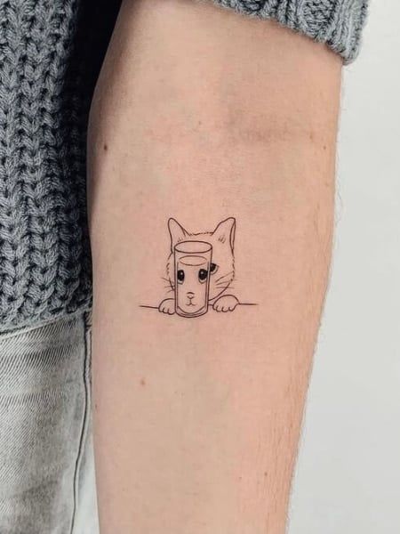 Minimalist cat tattoo peeking around a glass on forearm, wearing a knitted sweater.