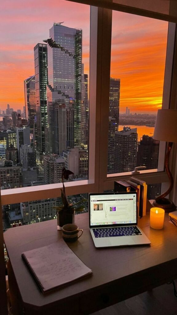 Home office view with a sunset over a city skyline, laptop, and candle on a desk.