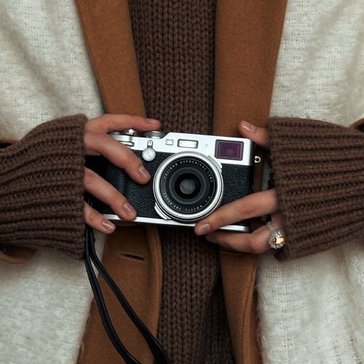 Person holding vintage camera, wearing brown sweater and coat, showcasing retro photography style.