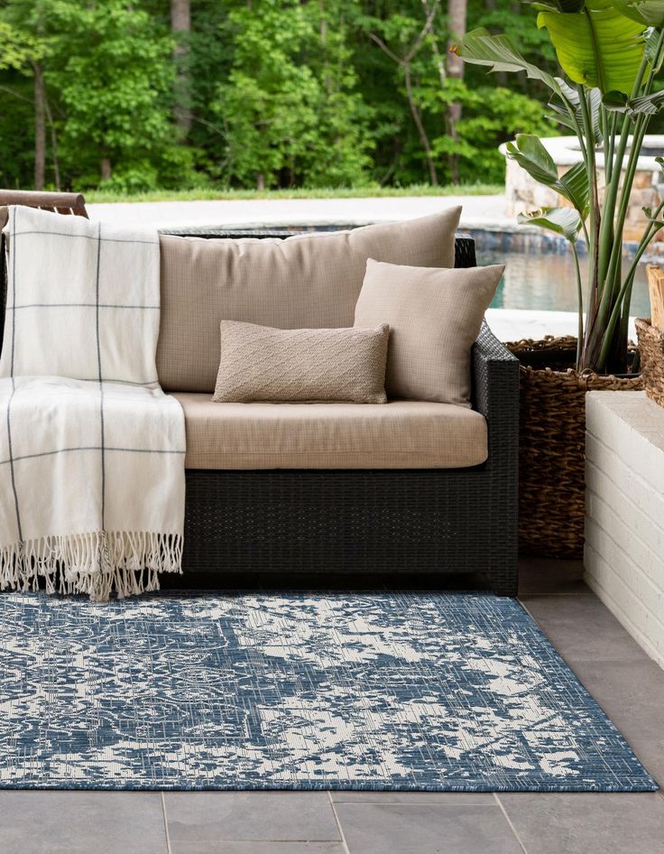 Cozy outdoor patio with beige sofa, decorative cushions, and blue patterned rug by a pool, surrounded by greenery.