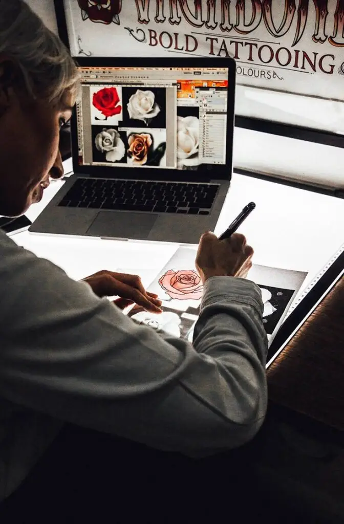 Tattoo artist designing rose tattoo with digital references on a laptop near a lightbox.