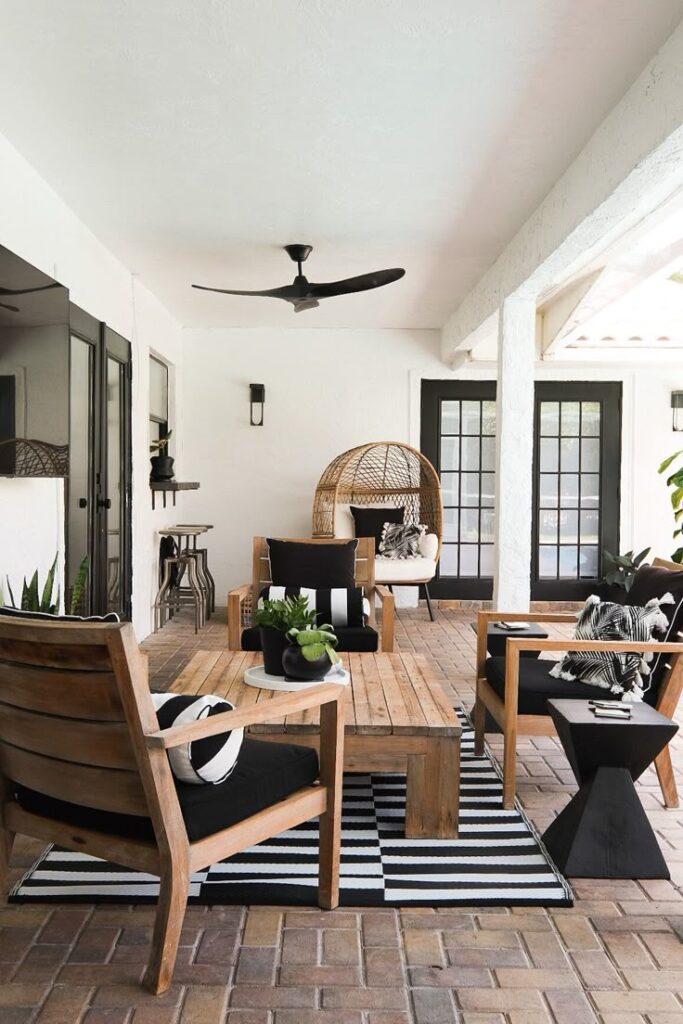 Modern patio with wooden furniture, black cushions, striped rug, and ceiling fan in a bright, cozy outdoor setting.