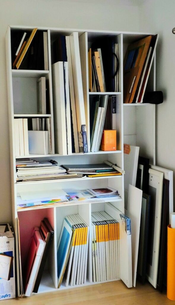 Organized art supplies on shelves, including canvases, sketchbooks, and paper, in a well-lit studio space.