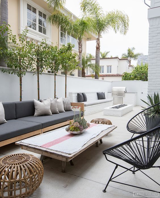 Modern outdoor patio with gray sofas, fire pit, and plants, set in a stylish backyard space with white walls.