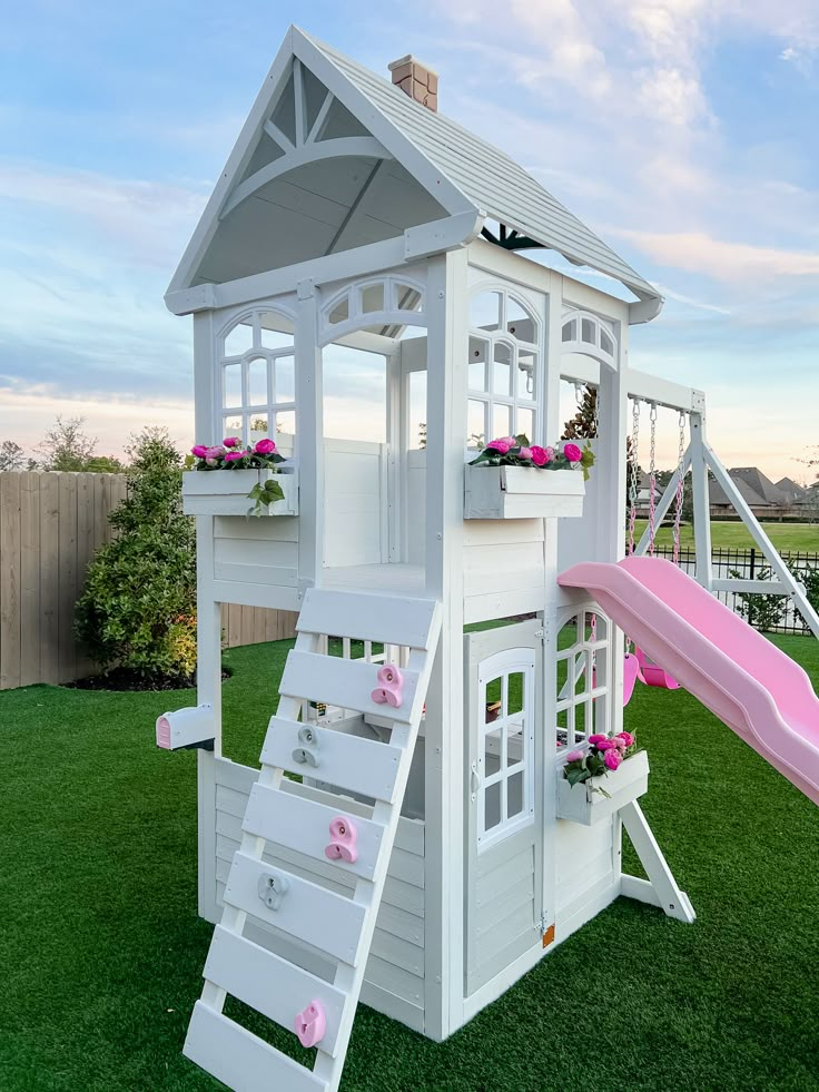 White playhouse with pink slide and climbing wall on green lawn, decorated with flowers.