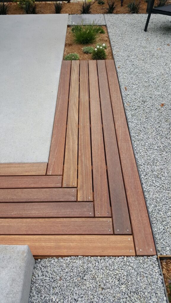 Modern garden design featuring wood decking, grey concrete, and gravel paths with planted greenery.