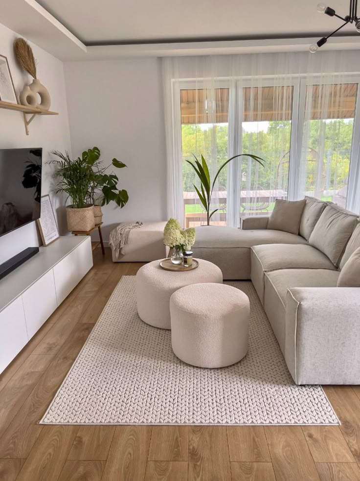 Modern living room with beige sectional sofa, round ottomans, plants, and large windows. Cozy and stylish interior design.