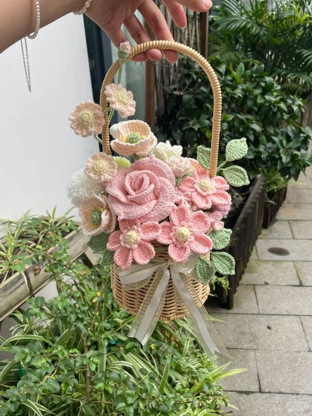 Hand holding a crocheted flower basket with pink, white blooms and green leaves, set against a garden backdrop.